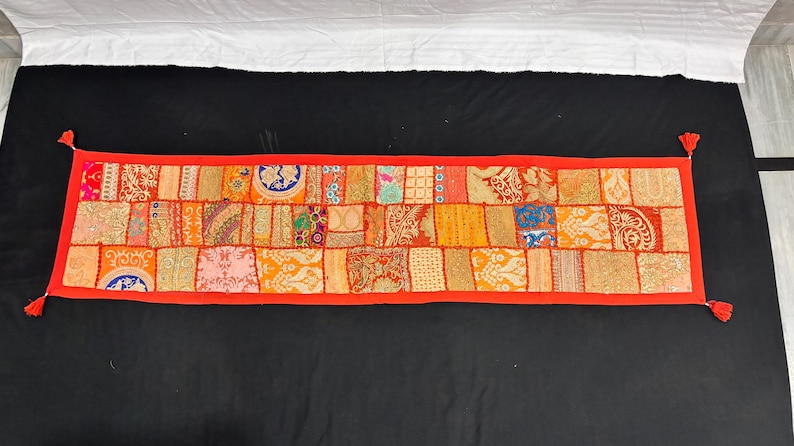 May include: A rectangular, patchwork textile in shades of orange, red, and gold. The design features a variety of patterned fabric squares, framed by a red border. Small red tassels are at each corner. This decorative piece is likely a table runner.