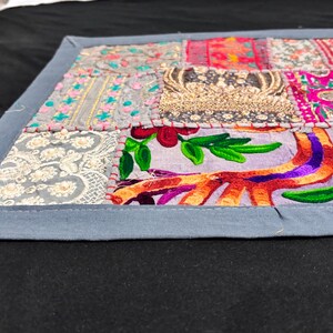 May include: A rectangular patchwork textile with a grey border. The patchwork features various fabric squares in shades of pink, purple, gold, and grey, with embroidered and beaded details. The textile is placed on a black surface.