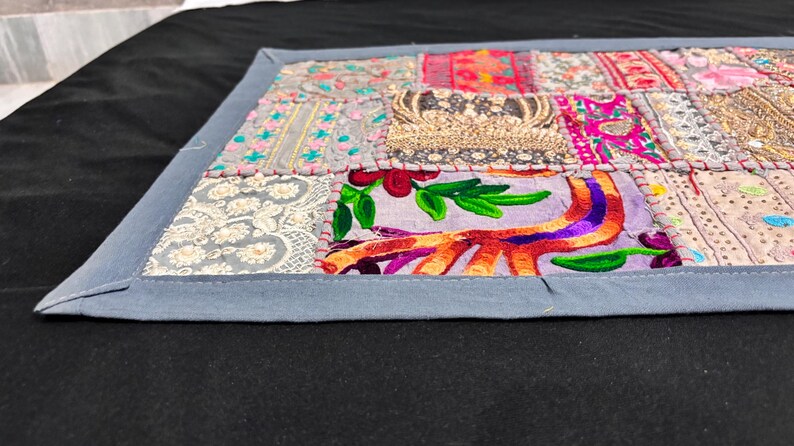 May include: A rectangular patchwork textile with a grey border. The patchwork features various fabric squares in shades of pink, purple, gold, and grey, with embroidered and beaded details. The textile is placed on a black surface.