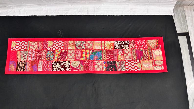 May include: A rectangular, patchwork textile with a vibrant red border. The interior is composed of numerous squares, each featuring a unique pattern in shades of red, pink, and gold. The textile is likely a decorative item, such as a table runner or wall hanging.