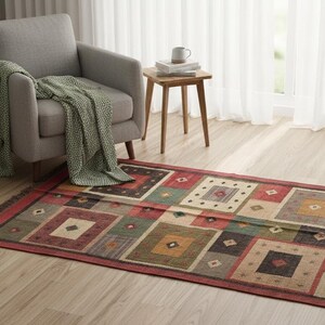 Könnte beinhalten: Ein rechteckiger Teppich mit geometrischem Muster in Rot-, Grün- und Beigetönen. Der Teppich hat ein Muster aus Quadraten und Rauten mit einem Fransenrand. Er liegt auf einem Holzboden, in der Nähe eines grauen Sessels und einer Topfpflanze.