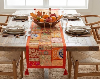 May include: A rustic wooden dining table set for a meal. The table is adorned with a colorful patchwork runner, a wooden bowl of fruit, and brass candle holders. Each place setting includes plates, silverware, and a glass of water. Woven chairs surround the table.