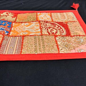 May include: A rectangular patchwork textile with a red border and tassels. The patchwork features various patterned squares in shades of orange, gold, blue, and red. The textile is likely a decorative item, such as a table runner or wall hanging.