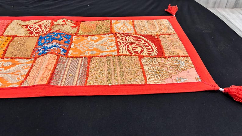 May include: A rectangular patchwork textile with a red border and tassels. The patchwork features various patterned squares in shades of orange, gold, blue, and red. The textile is likely a decorative item, such as a table runner or wall hanging.