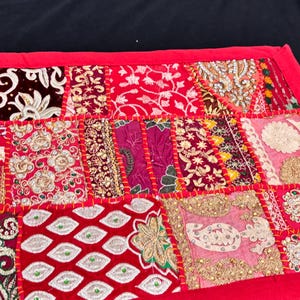 May include: A patchwork textile with a red border. The design features a variety of patterned squares in shades of red, pink, gold, and cream. The patterns include floral, paisley, and geometric designs, stitched together with red thread.
