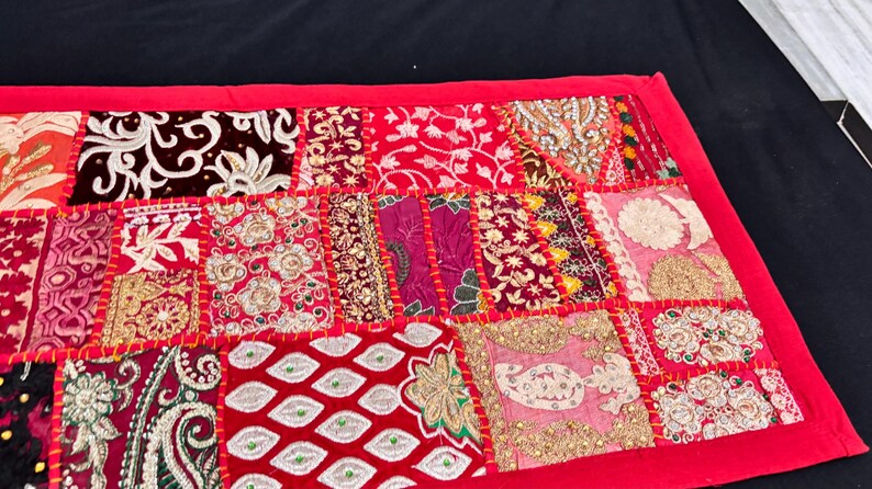May include: A patchwork textile with a red border. The design features a variety of patterned squares in shades of red, pink, gold, and cream. The patterns include floral, paisley, and geometric designs, stitched together with red thread.