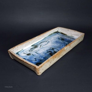 May include: Rectangular ceramic tray with a speckled, off-white rim and a marbled blue and white interior. The tray's interior features swirling patterns of blue and white, creating a unique, artistic design. The tray is a handmade piece.