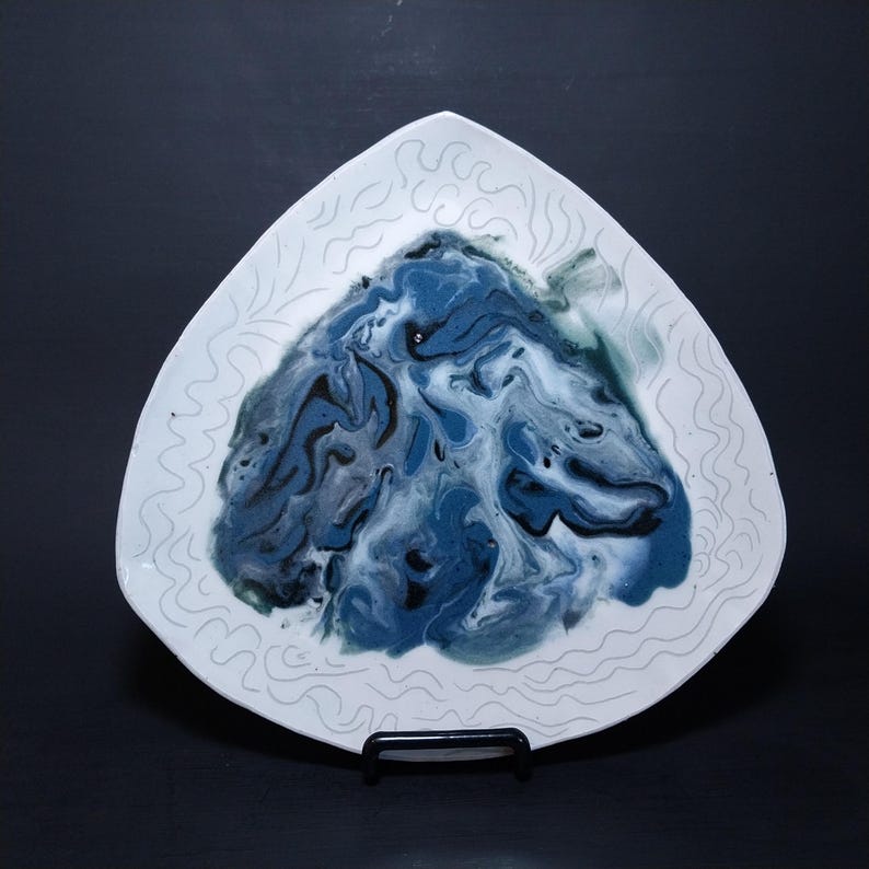 K&ouml;nnte beinhalten: Ein wei&szlig;er dreieckiger Keramikteller mit einem blauen, schwarzen und wei&szlig;en Marmormuster in der Mitte. Der Teller hat ein wei&szlig;es, wellenf&ouml;rmiges, gepr&auml;gtes Muster am Rand.