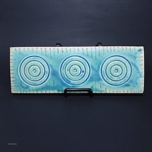 May include: A rectangular, turquoise ceramic tile with three concentric circle designs in varying shades of blue. The tile has a textured surface and is displayed on a black metal stand against a dark background. The edges feature a decorative pattern.