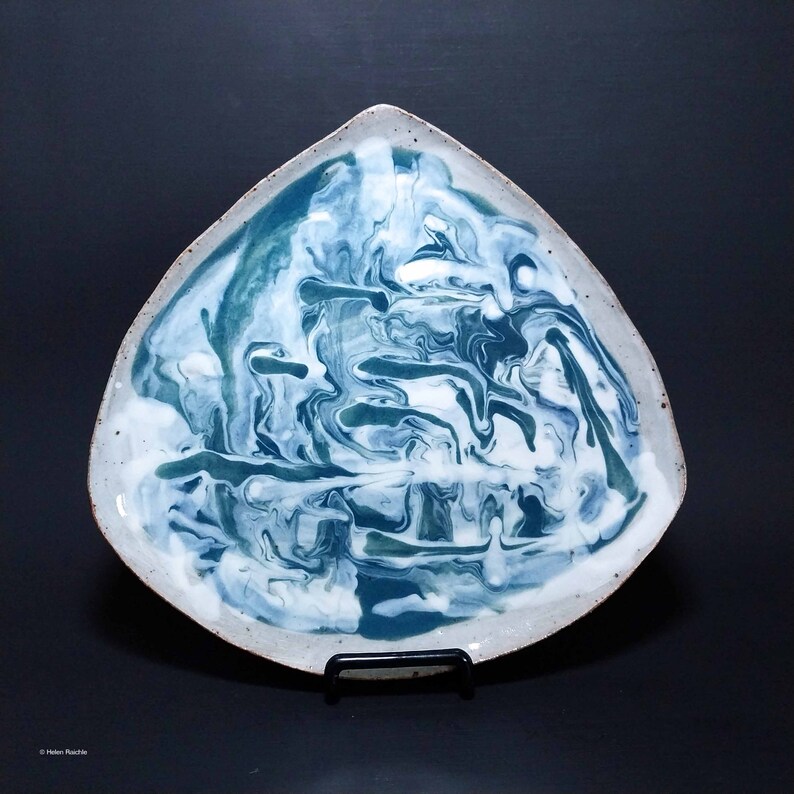 K&ouml;nnte beinhalten: Eine dreieckige Servierplatte aus Keramik mit einer wei&szlig;en und blauen Marmorierung. Die Platte hat eine leicht unregelm&auml;&szlig;ige Form und eine raue Textur.