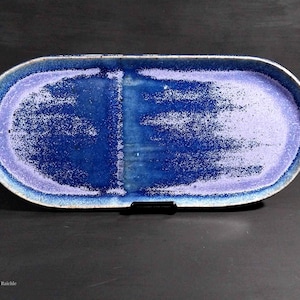 May include: An oval ceramic tray featuring a vibrant gradient glaze that transitions from deep royal blue in the center to a lighter lavender and white at the edges. The surface has a speckled texture with a distinct vertical band down the middle.