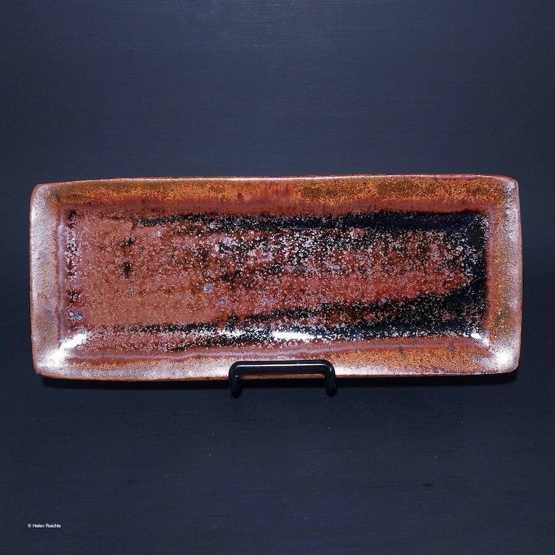 Puede incluir: Una bandeja rectangular de cer&aacute;mica con un esmalte moteado marr&oacute;n, negro y blanco. La bandeja tiene un borde ligeramente elevado y se muestra en un soporte negro.