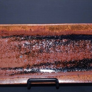 Puede incluir: Una bandeja rectangular de cer&aacute;mica con un esmalte marr&oacute;n, negro y plateado. La bandeja tiene una superficie texturizada y moteada.