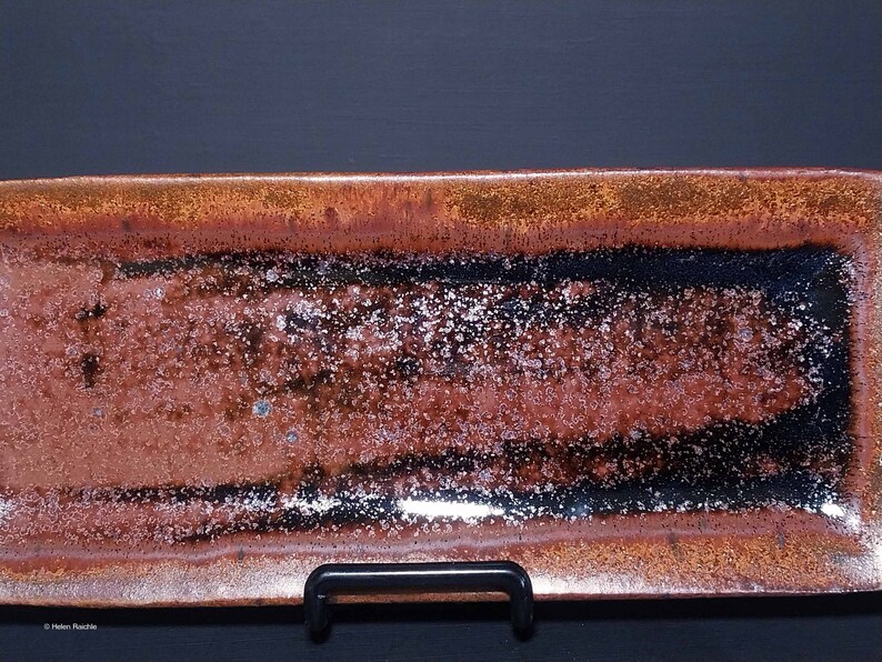 Puede incluir: Una bandeja rectangular de cer&aacute;mica con un esmalte marr&oacute;n, negro y plateado. La bandeja tiene una superficie texturizada y moteada.