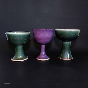 May include: Three ceramic chalices in shades of green and purple. The chalices have a pedestal base and a wide bowl. The chalice in the center is a vibrant purple color. The other two chalices are green.