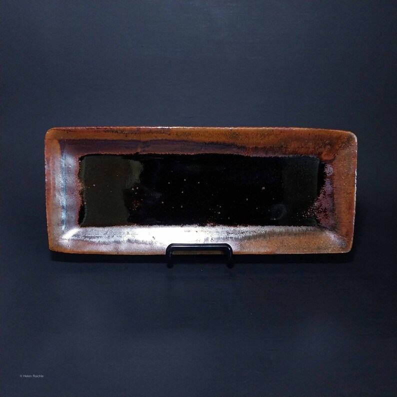 Puede incluir: Una bandeja rectangular de cer&aacute;mica con un esmalte marr&oacute;n oscuro y un brillo met&aacute;lico. La bandeja est&aacute; sobre un soporte negro.