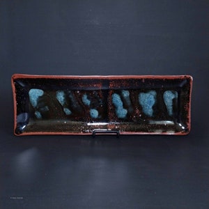 Puede incluir: Una bandeja rectangular de cerámica con un esmalte marrón oscuro y detalles azules. La bandeja tiene un acabado moteado y es perfecta para servir aperitivos o snacks.