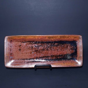 Puede incluir: Una bandeja rectangular de cer&aacute;mica con un esmalte marr&oacute;n y negro. La bandeja tiene un borde ligeramente elevado y est&aacute; sobre un soporte negro.