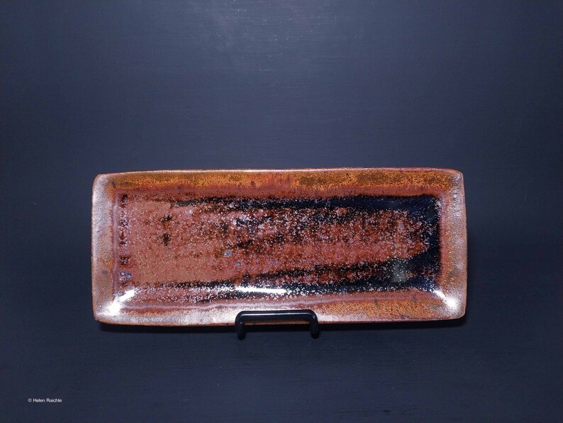 Puede incluir: Una bandeja rectangular de cer&aacute;mica con un esmalte marr&oacute;n y negro. La bandeja tiene un borde ligeramente elevado y est&aacute; sobre un soporte negro.