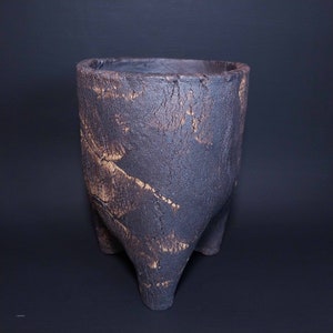 Pode incluir: Um vaso de cerâmica marrom escuro com uma superfície texturizada e três pernas. O vaso tem um aspecto rústico e terroso.