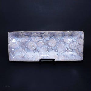 Puede incluir: Una bandeja rectangular de cerámica blanca con un diseño texturizado de guijarros. La bandeja es rectangular y tiene una ligera curva en los bordes.