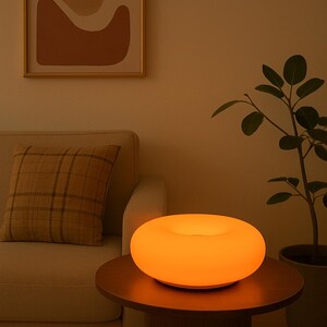 Peut inclure: Une lampe de table moderne en forme de donut, émettant une lueur orange chaude. La lampe est posée sur une table en bois brun foncé, avec un canapé beige et une œuvre d'art abstraite encadrée en arrière-plan. Une plante en pot ajoute de la verdure.