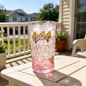 Op de afbeelding: Een hoog, helder glas gevuld met roze vloeistof en ijs. Het glas is versierd met een bloemenontwerp in roze, goud en wit. Het glas staat op een witte tafel op een zonnig terras. Ongeveer 15 cm hoog.