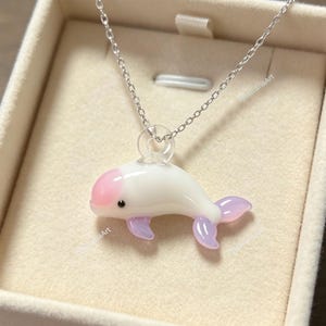 May include: A silver-toned chain necklace featuring a small, smooth, white dolphin-shaped pendant. The dolphin has a light pink patch on its head, a tiny black eye, and translucent light purple fins and tail. The pendant hangs from a clear loop.