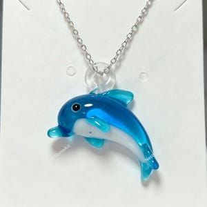 May include: A necklace featuring a blue and white glass dolphin pendant suspended from a silver-toned metal chain. The dolphin is depicted in a swimming pose with a clear glass loop at the top for the chain to pass through, showcasing a smooth finish.