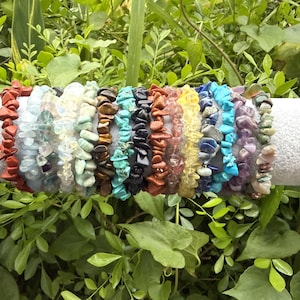 May include: A collection of colorful gemstone chip bracelets displayed on a white cylindrical surface. The bracelets feature various colors, including red, blue, green, and yellow, and are set against a backdrop of lush green foliage.
