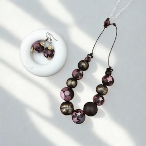 Op de afbeelding: Een ketting en oorbellen set. De ketting heeft grote, ronde kralen in bruin, goud en roze, sommige met sterpatronen. De oorbellen hebben vergelijkbare kralen met sterpatroon. De sieraden worden gepresenteerd op een wit oppervlak.