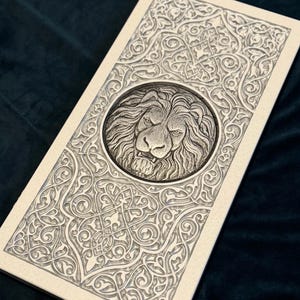 May include: An off-white rectangular plaque with an embossed lion's head in a circular frame. The plaque is decorated with detailed, swirling patterns around the lion and on the borders. The design is in a light grey colour.