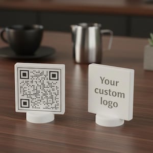 Könnte beinhalten: Zwei weiße Tischschilder, eines mit einem QR-Code und das andere mit dem Text "Your custom logo". Beide Schilder stehen auf einer dunklen Holzoberfläche. Eine schwarze Kaffeetasse und ein Edelstahlkrug sind im Hintergrund.