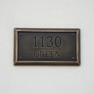 May include: Rectangular address plaque with the numbers "1130" and the word "GREEN" in a bronze finish. The plaque is framed with a decorative border and mounted on a white textured wall.