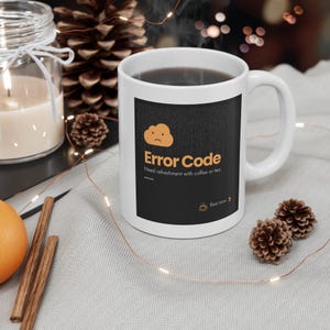 May include: White ceramic mug with a black graphic that reads "Error Code" and "Need refreshment with coffee or tea." The mug is filled with a dark liquid and sits on a light gray surface with decorative elements.