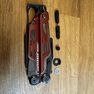 Könnte beinhalten: Ein rotes und schwarzes Leatherman Signal Multitool mit dem Markennamen auf der Seite. Das Werkzeug ist geöffnet und zeigt verschiedene Komponenten. Mehrere schwarze und silberne Zubehörteile liegen neben dem Werkzeug.