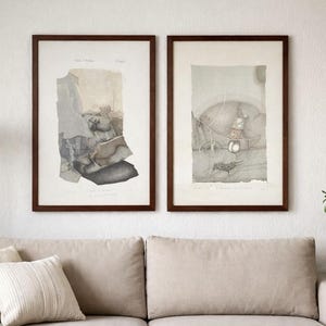 Op de afbeelding: Twee ingelijste kunstdrukken met donkerbruine frames hangen boven een beige bank. De prints tonen abstracte, gedempte kleurenillustraties. Een witte vloerlamp en een plant in een pot zijn ook zichtbaar.