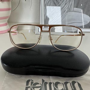 Könnte beinhalten: Goldgerahmte Brille mit rechteckiger Form, auf einem schwarzen ovalen Etui platziert. Die Gläser sind klar, und das Design umfasst einen Doppelsteg. Ein Reinigungstuch mit der Marke "felmann" ist sichtbar.