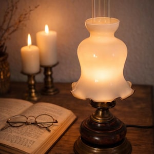 Könnte beinhalten: Eine Tischlampe im Vintage-Stil mit weißem, gerüschtem Schirm und einem transparenten Glaszylinder. Die Lampe hat einen dunklen Holzsockel mit Messingakzenten. Zwei brennende Kerzen und ein offenes Buch mit Brille sind ebenfalls zu sehen.