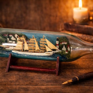 Puede incluir: Maqueta detallada de un velero dentro de una botella de vidrio, apoyada sobre un soporte de madera. El barco tiene múltiples velas y está ambientado en un paisaje costero. También se ven un bolígrafo y una vela.