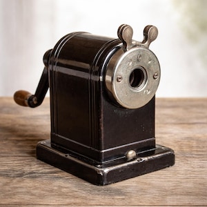 Könnte beinhalten: Ein schwarzer Bleistiftanspitzer im Vintage-Stil mit einer silbernen Metallfront und einem Holzgriff. Der Anspitzer hat eine rechteckige Basis und eine Kurbel an der Seite. Das Design ist klassisch und funktionell.