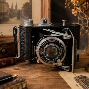 May include: A vintage black folding camera with a silver Compur lens assembly. The lens features the text Welta, 1:2.9, f=7.5cm, and Compur. The camera body has a textured black leatherette finish, a silver winding knob, and a metal viewfinder.