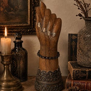 May include: A wooden hand sculpture with silver-painted fingernails and a decorative metal base. A dark bracelet encircles the wrist. The sculpture is displayed with a candle, antique books, and a vase, creating a vintage aesthetic.