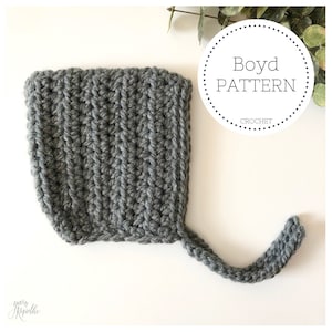 Könnte beinhalten: Graue gehäkelte Baby-Mütze mit langem Band. Die Mütze ist mit einem einfachen Stichmuster gearbeitet und hat einen klassischen Look. Die Mütze ist mit "Boyd PATTERN CROCHET" beschriftet.