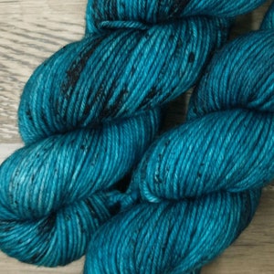 May include: Two skeins of teal-colored yarn with black speckles. Each skein has a white label with the text "ATIC WOOLS" and a sheep logo. The yarn is arranged on a light brown wooden surface.