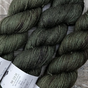 May include: Three skeins of dark green yarn with white labels that read "AJHC WOOLS" and "Yak Silk DK". The yarn is a deep olive color, with subtle variations in tone. The skeins are arranged on a light wood surface.