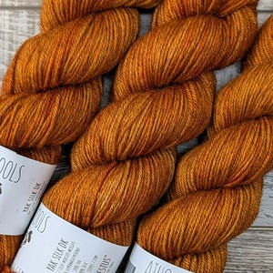 May include: Three skeins of orange-brown yarn with white labels that read "ATHC WOOLS" and "Yak Silk DK". The yarn is a rich, warm color, perfect for knitting or crocheting projects. The labels also feature a sheep logo and the text "HERPHAESTUS".