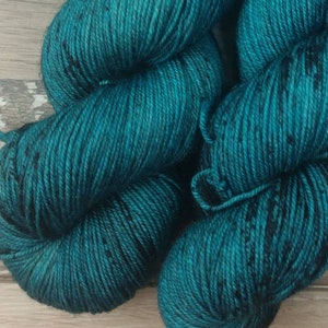 May include: Two skeins of teal-colored yarn with black flecks. The yarn is tightly wound and has a soft, textured appearance. White labels with the text "ATHC WOOLS" and a sheep logo are visible.