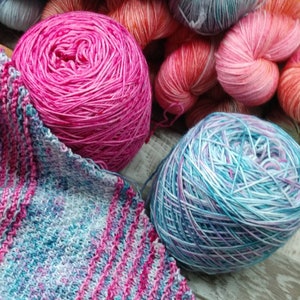 May include: A close-up of three balls of yarn in pink, blue, and white. The yarn is arranged on a wooden surface.