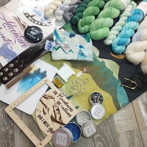 May include: A collection of knitting and crafting supplies, including skeins of yarn in various shades of green, blue, and white. The image also features stitch markers, beads, a leather cuff, and a wooden frame with the text "The Mountains are calling and I must go."